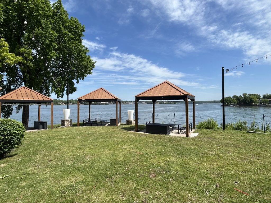 Three open-air gazebos with metal roofs overlook Lake of the Ozarks at Lazy Gators in Missouri, set on a grassy lawn with string lights above and calm blue water in the background on a sunny day.
