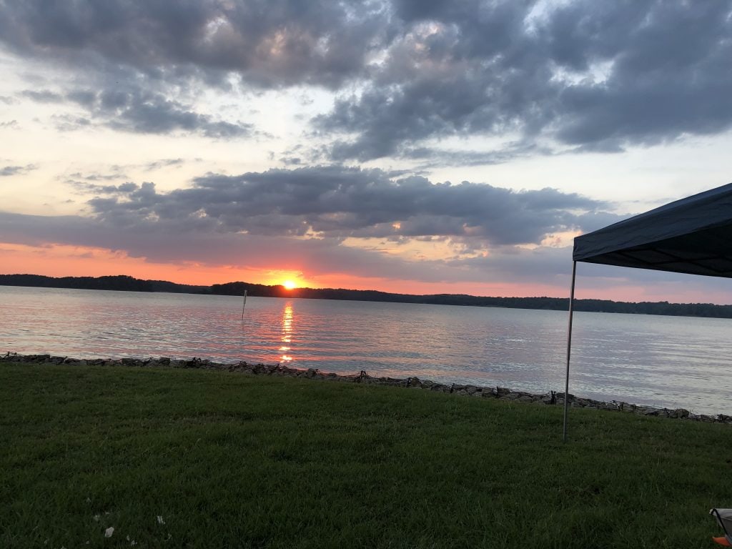 A vibrant sunset over Lake Lanier casts warm orange and pink reflections on the water, viewed from a grassy shoreline at Old Federal Campground. A pop-up canopy is partially visible on the right, suggesting a relaxed evening setup by the lake.
