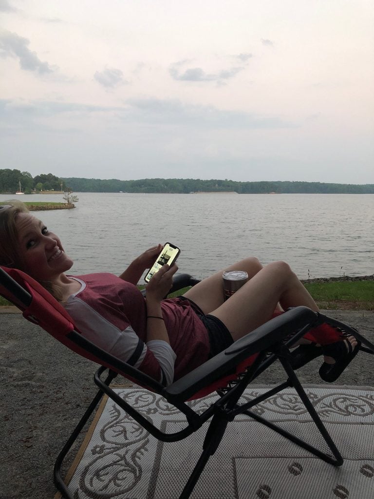 A woman relaxes in a reclining camp chair at Old Federal Campground, smiling while looking at her phone with a drink by her side. Behind her, Lake Lanier stretches out under an overcast sky, creating a calm, peaceful atmosphere.