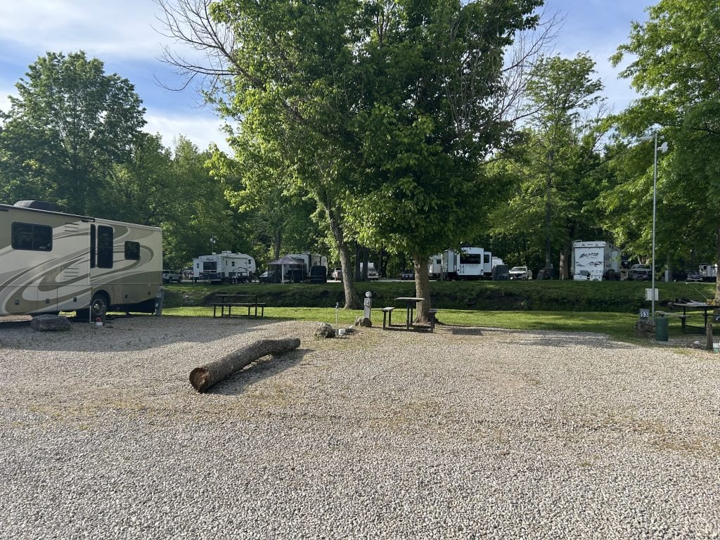 A gravel RV campsite with picnic tables and a fire ring sits empty under sunny skies, with a large log lying on the ground in the foreground. Several motorhomes and fifth-wheel RVs are parked in the background among lush green trees.