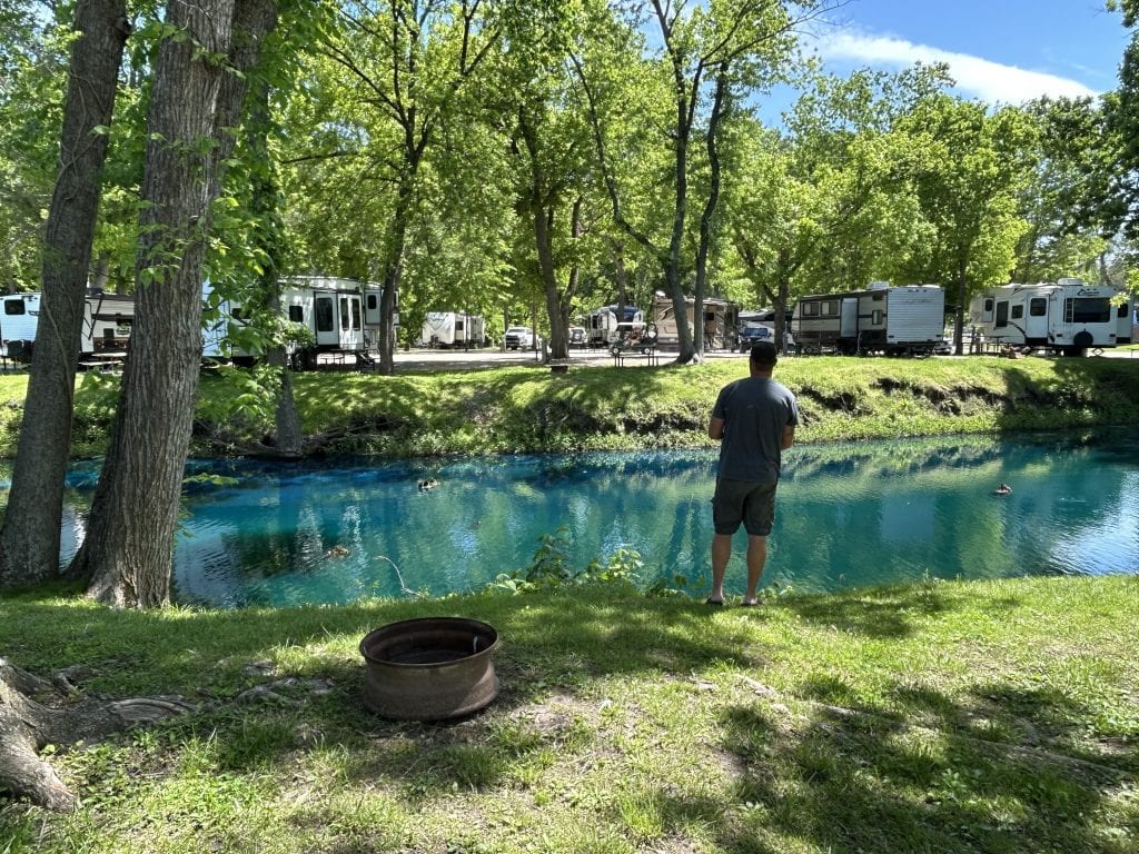Alan stands barefoot at the edge of a vibrant blue pond, fishing under the shade of tall trees at Ozarks Landing RV Resort in Missouri. Several RVs are parked across the pond beneath the leafy canopy, creating a peaceful campground setting on a sunny day.