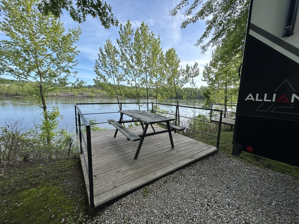 A wooden deck with a wood and metal picnic table and metal railings overlooks a calm river at Ozarks Landing RV Resort in Missouri, surrounded by leafy green trees and positioned beside a parked Alliance RV.