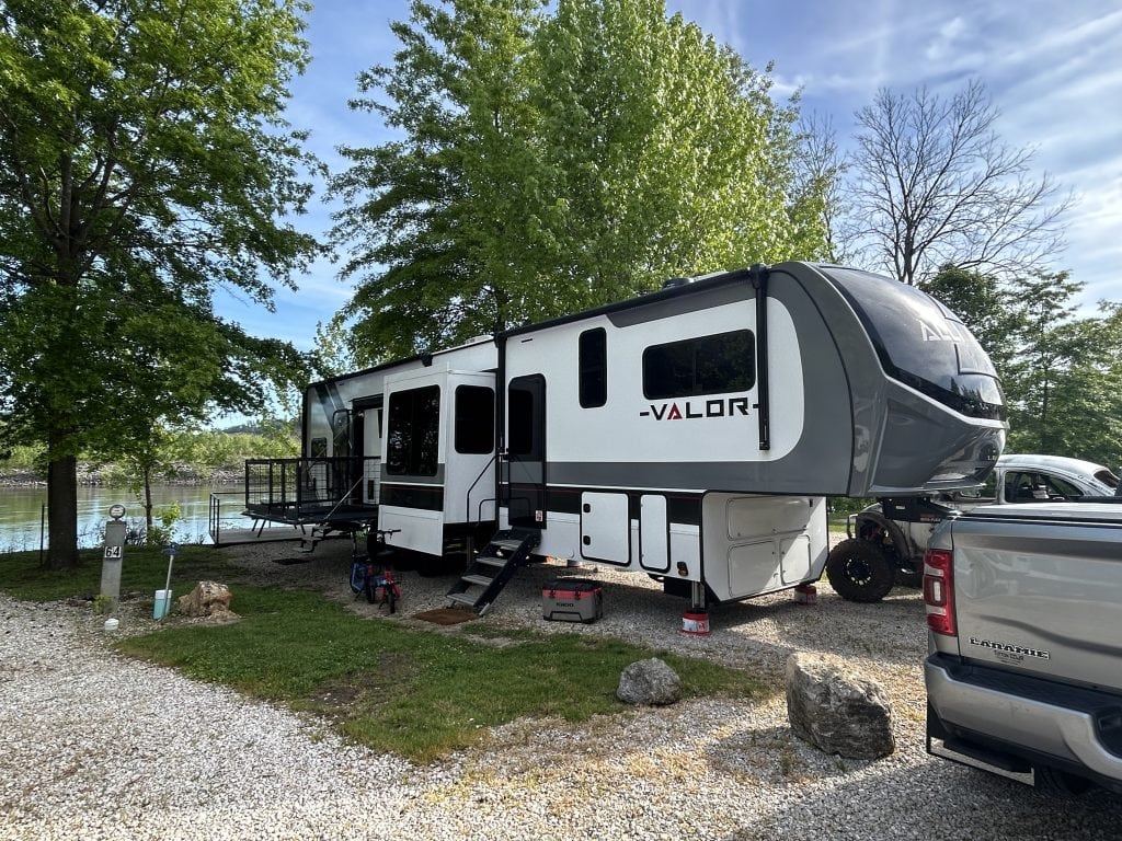 A large Alliance Valor fifth-wheel RV is parked at a lakeside campsite, connected to a silver Ram truck. The RV has an outdoor patio deck extended over the water-facing side, with trees providing shade and the lake visible in the background.