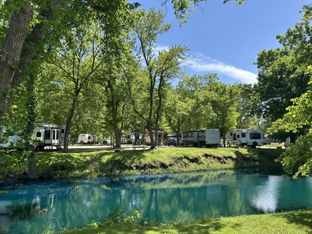 Several RVs are parked under leafy green trees along a shaded campground road at Ozarks Landing RV Resort in Missouri, with a bright blue pond in the foreground reflecting the clear summer sky.