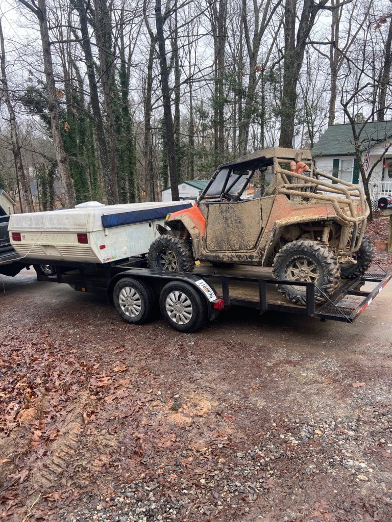 A mud-covered orange and black side-by-side ATV sits on a trailer alongside a folded white pop-up camper. The trailer is parked on a wet, leaf-strewn driveway with bare winter trees and houses in the background.