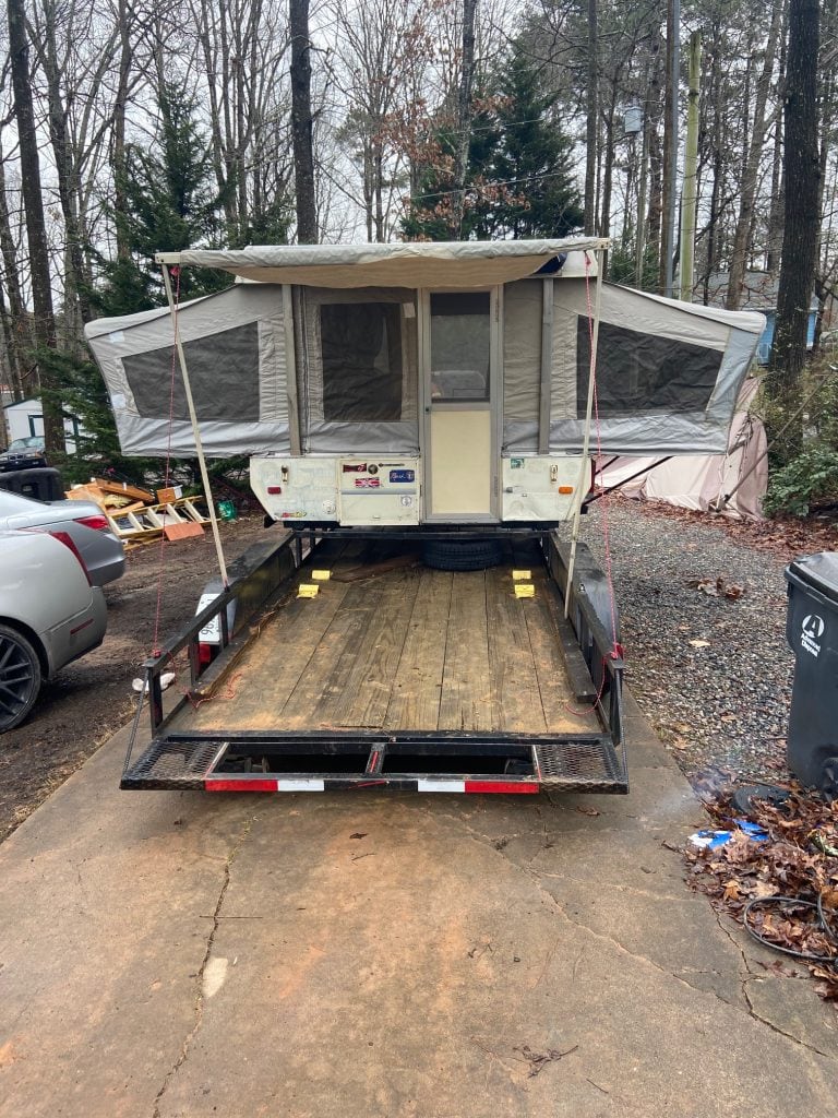 A small pop-up camper is set up on a utility trailer parked in a wooded driveway, with the sides extended and mesh windows visible under an overcast sky.
