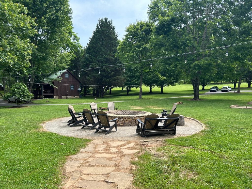A circular stone fire pit is surrounded by Adirondack chairs on a gravel pad, with string lights overhead. The setting is a grassy yard bordered by tall trees, with a rustic wooden cabin and parked cars in the background.