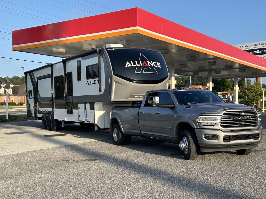 A large Valor fifth-wheel RV hitched to a gray Ram dually truck is parked at a gas station under a bright red and yellow canopy on a sunny day.