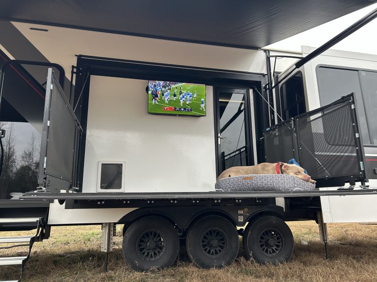 Coco, a large tan dog with a red collar, rests on a cushioned bed on the rear patio deck of a fifth-wheel RV. A TV mounted on the RV wall plays a football game, and the patio is enclosed with black safety railings under an extended awning.
