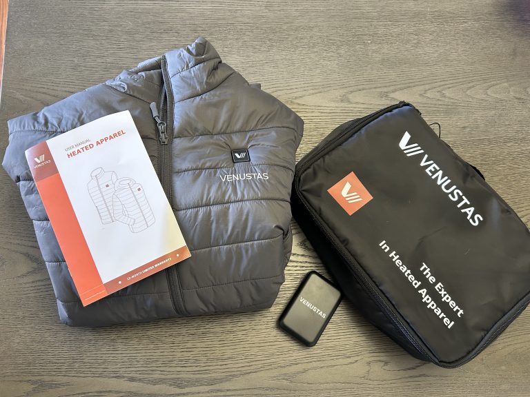 Flat lay of a Venustas heated vest set including the gray puffer-style vest, a black zippered storage bag labeled “The Expert in Heated Apparel,” a user manual with product illustrations, and a compact Venustas-branded battery pack. All items are displayed on a wood-grain surface.