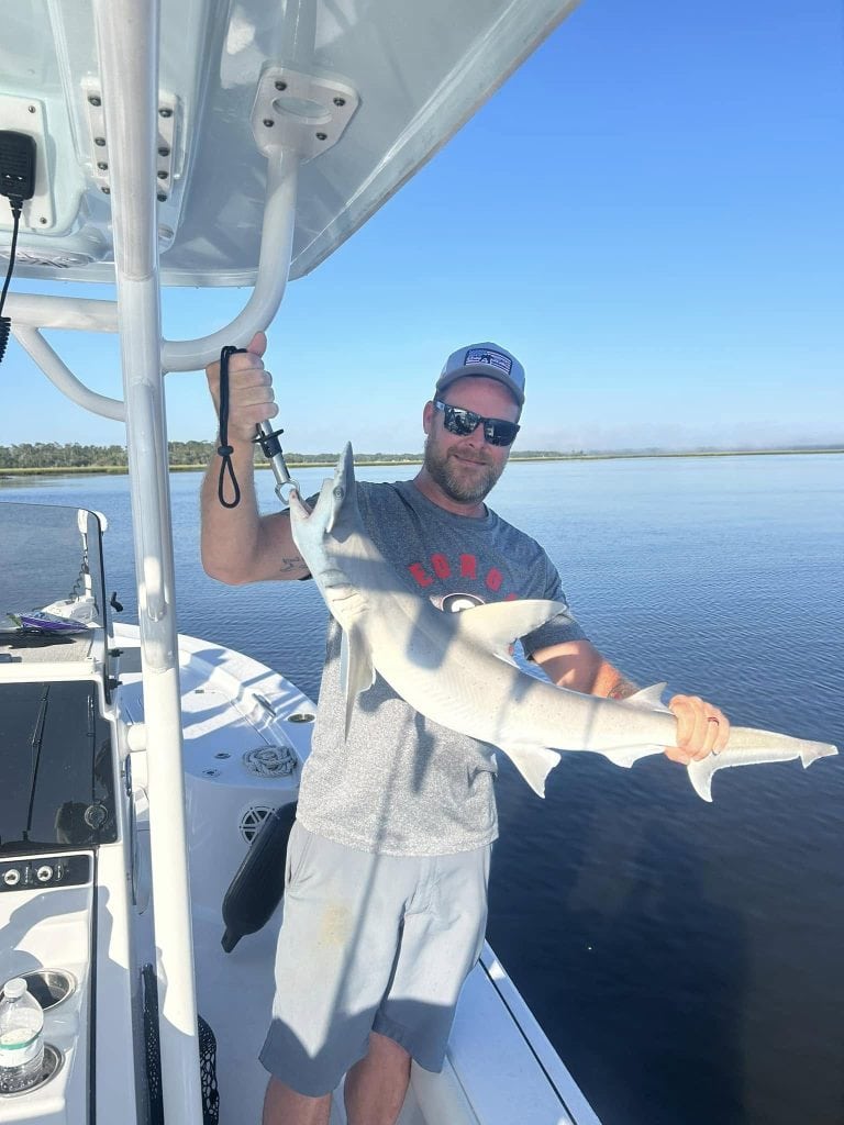 Alan standing on a fishing boat holding up a small shark with both hands, smiling at the camera under a clear blue sky. The calm waters and distant shoreline suggest a peaceful fishing trip near Amelia Island.