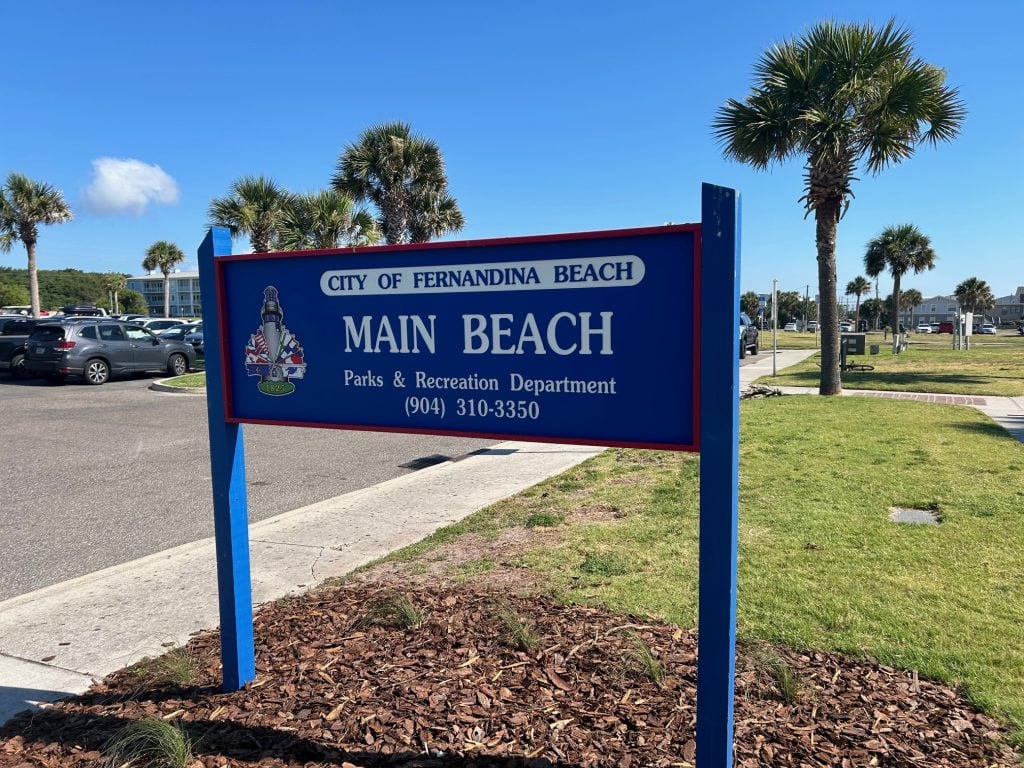 Blue and red sign for Main Beach in the City of Fernandina Beach, placed near palm trees, a grassy park, and a beachside parking lot under a clear blue sky. A popular public beach on Amelia Island managed by the Parks & Recreation Department.
