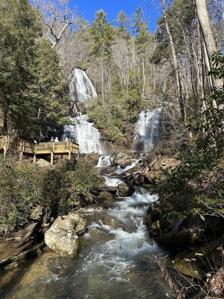 A bright winter view of Anna Ruby Falls shows twin streams of water cascading down a rocky cliff into a rushing creek, framed by evergreen and leafless trees under a clear blue sky. A wooden observation deck on the left provides visitors a closer look at the scenic waterfalls.