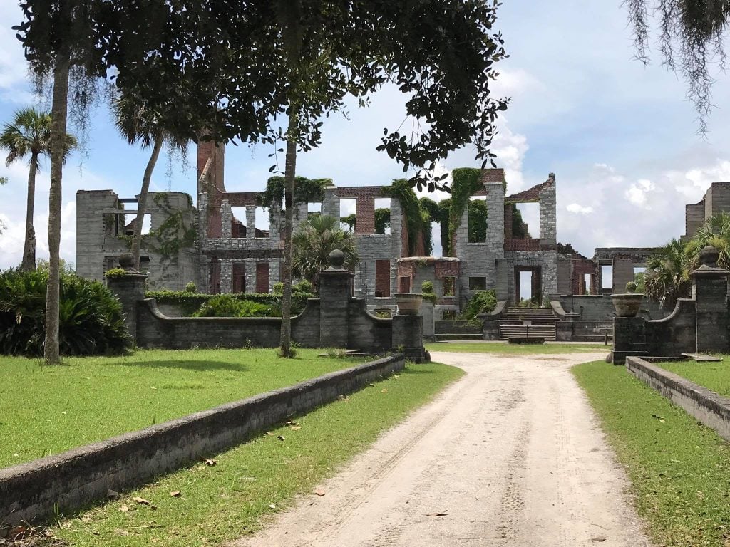 Sunlight filters through moss-draped oak trees framing the Dungeness Ruins, a large stone mansion with crumbling walls and empty windows on Cumberland Island. A sandy path leads to the historic site, surrounded by palm trees and green lawns.