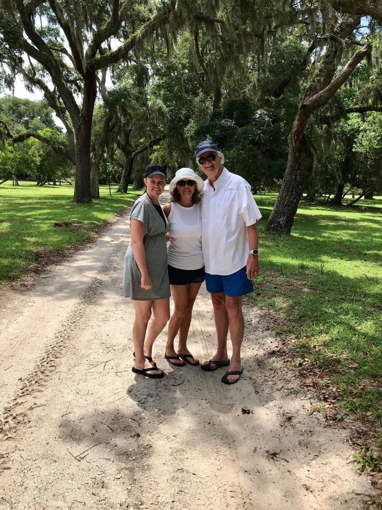 Three people pose together on a sandy path beneath large oak trees draped in Spanish moss on Cumberland Island. The lush green surroundings and dappled sunlight create a relaxed, scenic backdrop for their outdoor stroll.