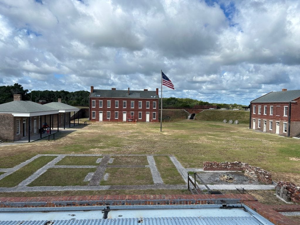 View of the parade ground at Fort Clinch, showing restored 19th-century red-brick military buildings, a large American flag at center, and outlines of former structures marked on the grass. The sky is partly cloudy, and the historic fort's defensive walls are visible in the background.