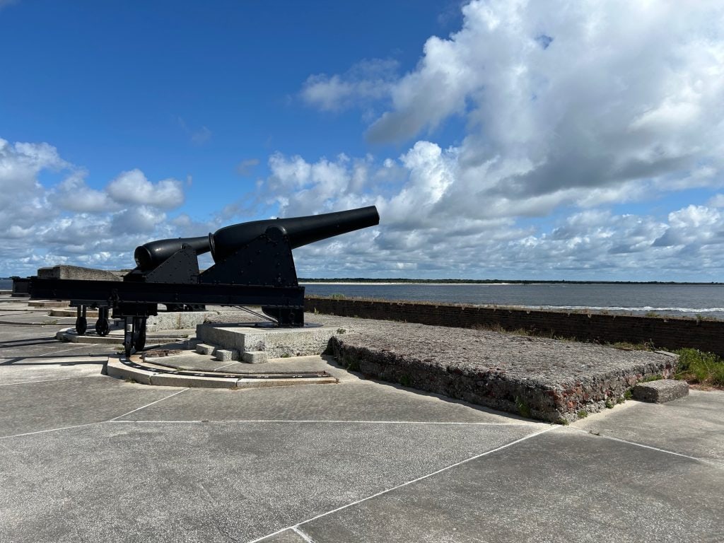 Historic black cannon mounted on a concrete platform at Fort Clinch, overlooking the water under a partly cloudy sky. The cannon faces out toward the coastline, reflecting the fort’s strategic position for coastal defense.