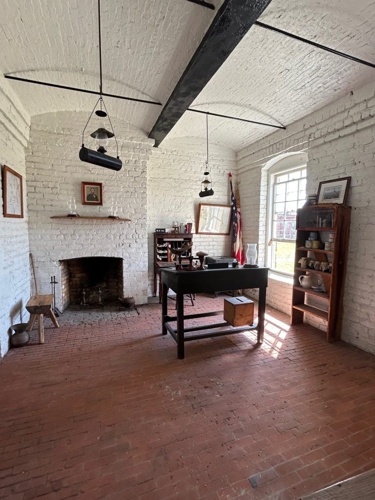 Historic officer's quarters at Fort Clinch with whitewashed brick walls, a brick fireplace, and period furnishings including a black wooden desk, shelves with supplies, and framed portraits. Sunlight streams through a multi-pane window, illuminating the preserved 19th-century room.