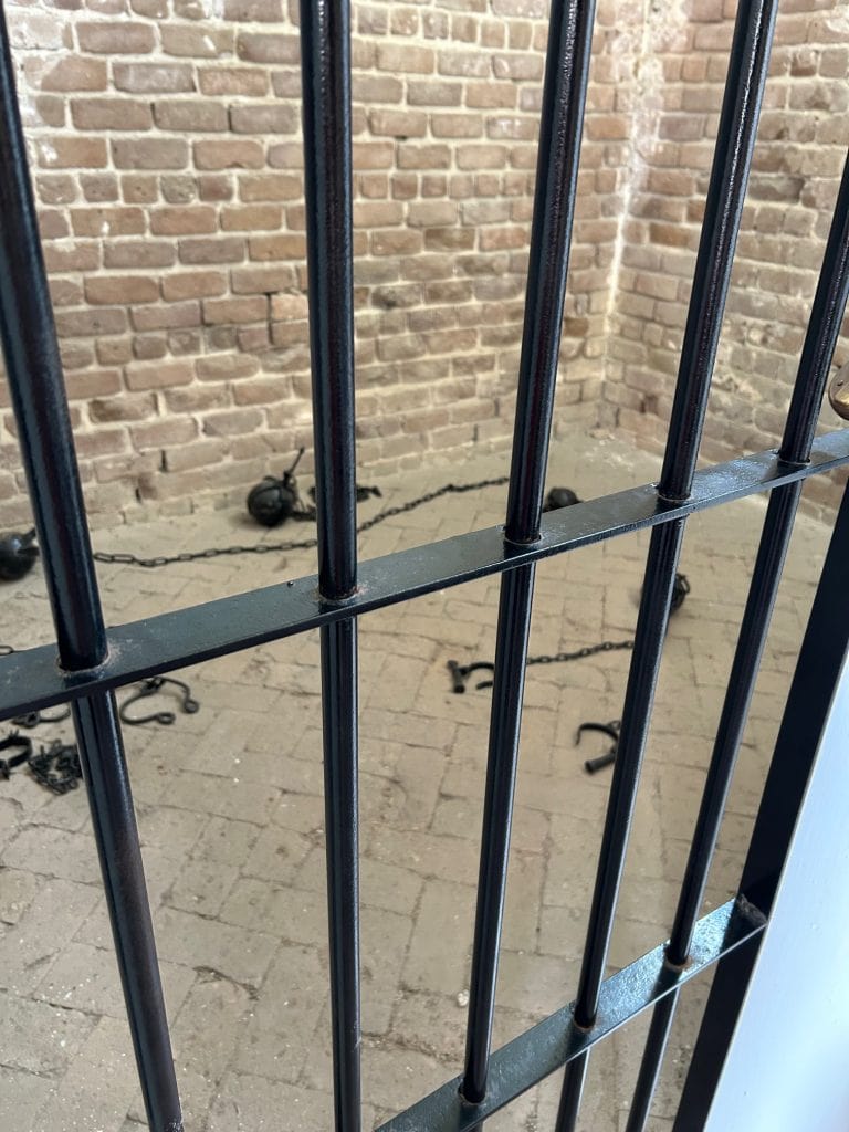 View through black metal bars into a historic brick prison cell at Fort Clinch, showing shackles and chains lying on the stone floor. The scene evokes the fort's 19th-century military prison history.