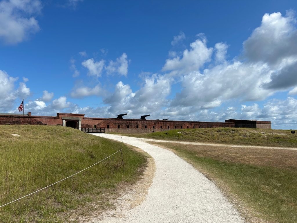 Gravel path leading to the entrance of Fort Clinch, a large red-brick Civil War-era fort with black cannons mounted along the top and an American flag flying above the gate. The surrounding grassy dunes and blue sky add to the historic coastal setting.