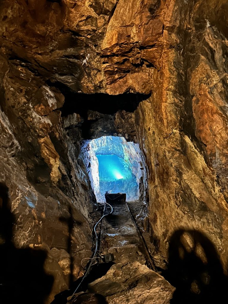 A narrow tunnel inside the Consolidated Gold Mine in Dahlonega leads to a glowing blue-lit chamber, with jagged rock walls and mining equipment casting shadows along the passage. Cables and lighting fixtures hint at the modern touches in this historic underground space.
