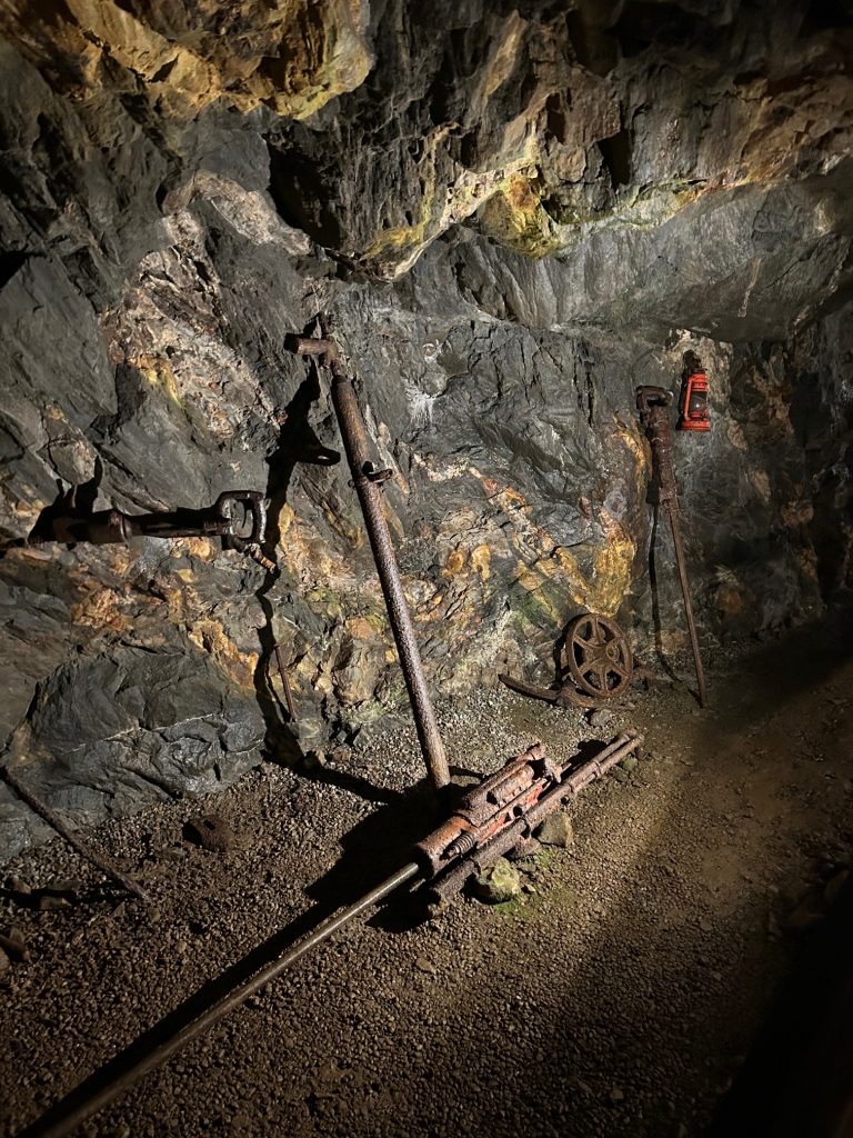Rusty vintage mining tools and a red lantern are displayed against the rock wall inside the Consolidated Gold Mine in Dahlonega. The equipment, partially embedded in the stone, shows the worn and rugged remnants of historic gold mining operations.