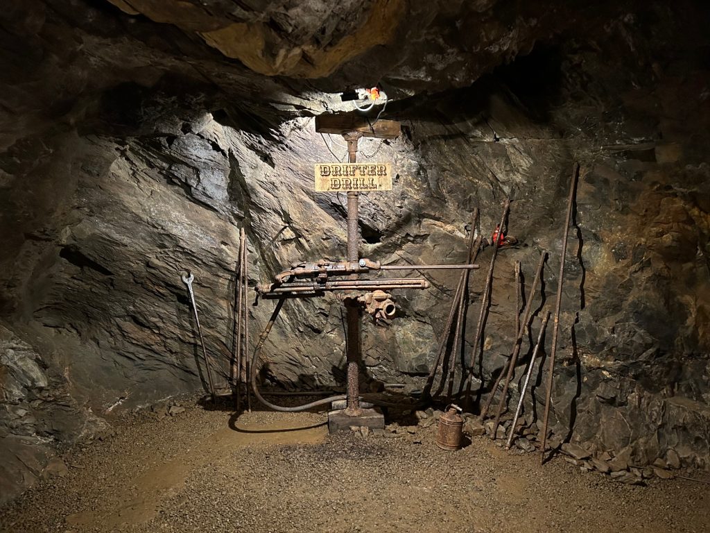 An old drifter drill display inside the Consolidated Gold Mine in Dahlonega features a mounted pneumatic drill surrounded by long metal drill rods, a wrench, and other vintage mining tools. A wooden sign labeled “Drifter Drill” hangs above the setup against the rocky tunnel wall.
