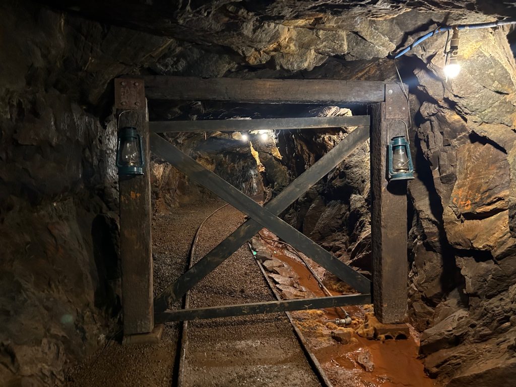Inside the Consolidated Gold Mine, a wooden support gate with two lanterns frames a narrow mining tunnel with rail tracks and damp, rust-colored ground. Overhead lighting illuminates the rugged rock walls and pathway deep underground.