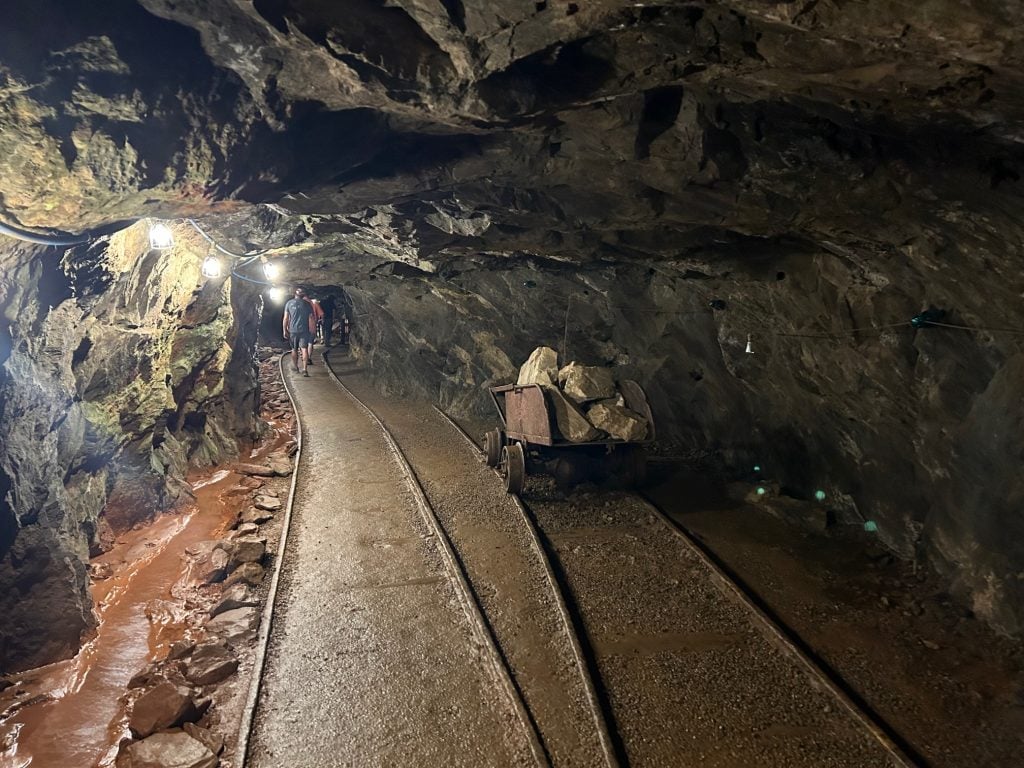 Visitors walk through a dimly lit tunnel inside the Consolidated Gold Mine in Dahlonega, passing a rusted mining cart filled with large rocks. Overhead lights illuminate the rough rock walls and a narrow water channel running alongside the tracks.