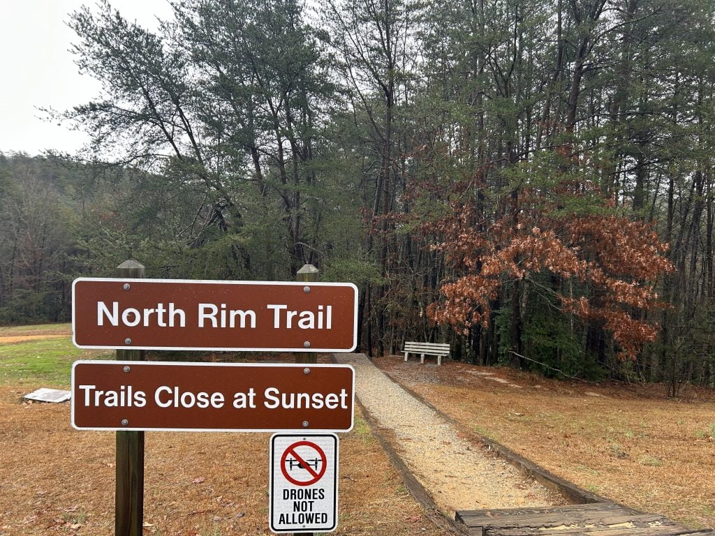 Trailhead sign for the North Rim Trail at Tallulah Gorge State Park, with additional notices stating “Trails Close at Sunset” and “Drones Not Allowed.” The gravel path leads into a wooded area with a white bench just inside the tree line.