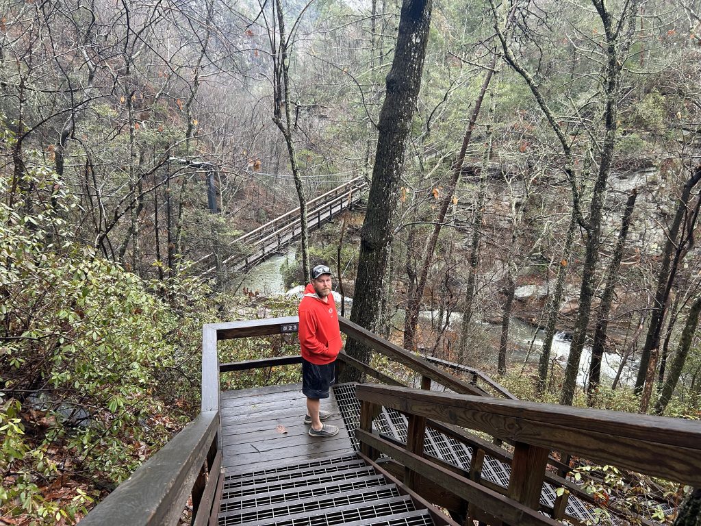 A man in a red hoodie stands on a wooden landing partway down the metal-grated stairs leading to the Tallulah Gorge suspension bridge. The bridge stretches over a rushing river below, framed by steep rocky cliffs and dense, leafless forest.