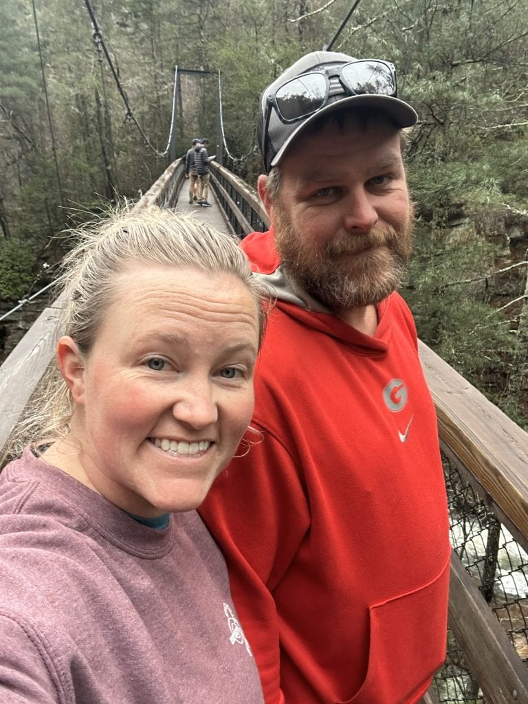 Two people smile for a selfie on the Tallulah Gorge suspension bridge, with forested cliffs and a few other hikers in the background. The wooden planks and steel cables of the bridge frame the scene, capturing the adventure and natural beauty of the hike.