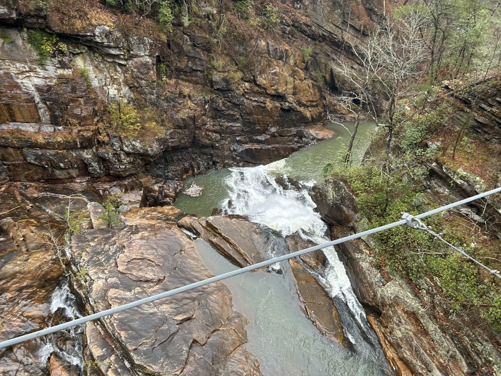 View from the Tallulah Gorge suspension bridge looking down at a cascading waterfall cutting through layered, rust-colored rock formations. The rushing water flows into a narrow greenish pool below, surrounded by steep canyon walls and sparse vegetation.
