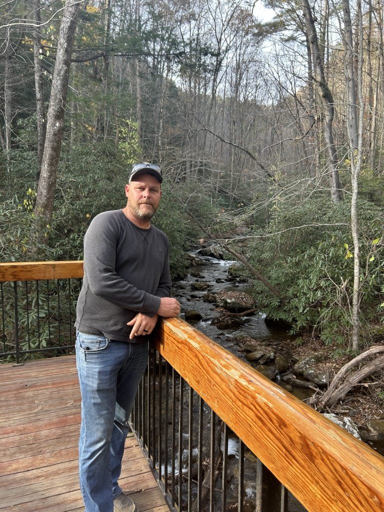 A man in a dark long-sleeve shirt, jeans, and a baseball cap leans against a wooden railing on a viewing platform above a rocky creek, surrounded by dense forest near Anna Ruby Falls. Bare trees and evergreen shrubs line the stream in a peaceful late autumn setting.