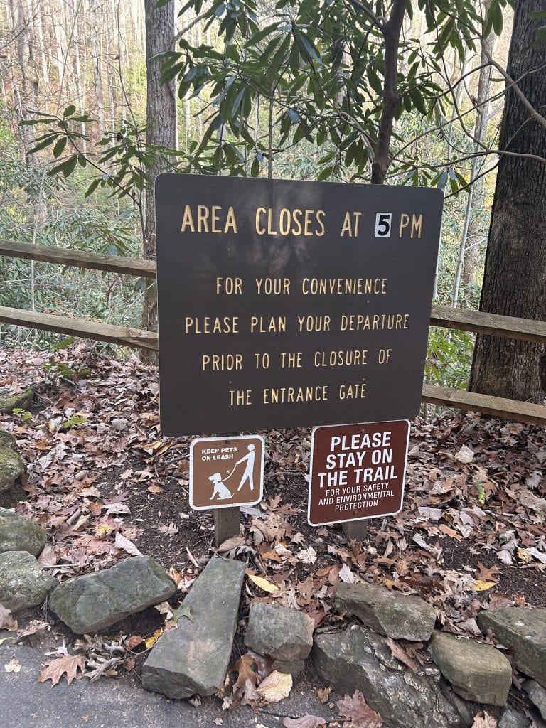 A posted sign along the Anna Ruby Falls trail reads “Area closes at 5 PM. For your convenience please plan your departure prior to the closure of the entrance gate.” Additional smaller signs below advise visitors to keep pets on a leash and to stay on the trail for safety and environmental protection.