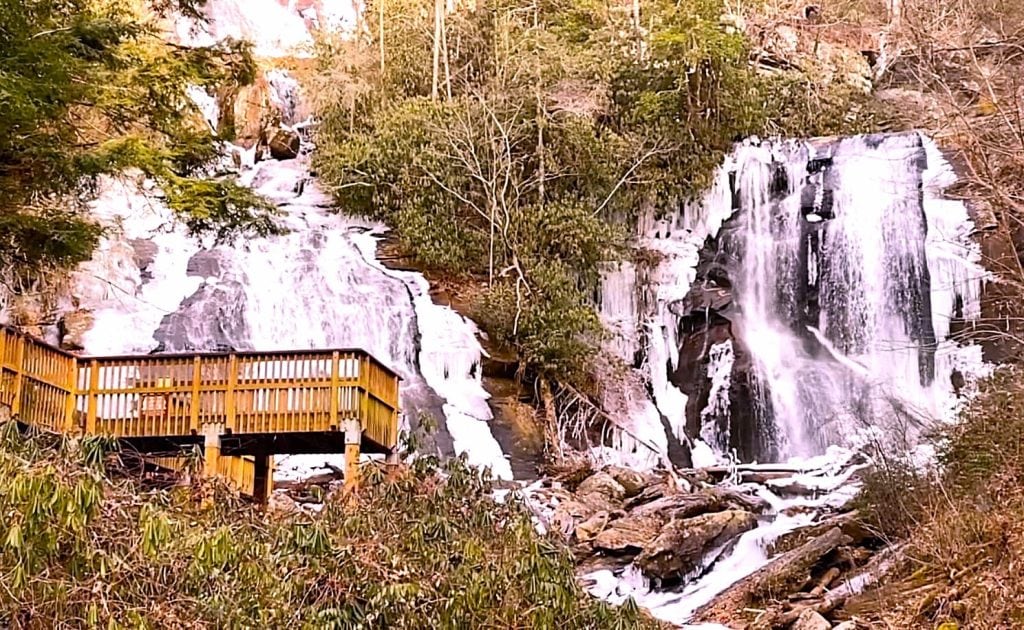 Twin streams of Anna Ruby Falls cascade down a rocky cliff surrounded by winter foliage and patches of ice, with a wooden observation deck in the foreground offering a close-up view of the frozen waterfall scene.