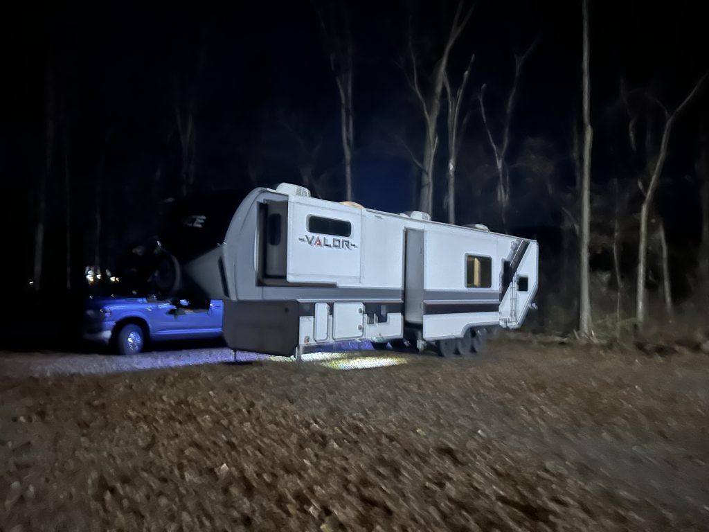 Fifth-wheel RV labeled "Valor" parked in a wooded area at night, hitched to a pickup truck with LED lights illuminating the ground beneath. Bare trees surround the campsite, creating a quiet, remote atmosphere ideal for a Harvest Hosts overnight stay.