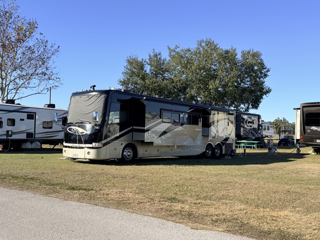 A beige and black Class A motorhome with slide-outs extended is parked on a grassy campsite next to other RVs under a clear blue sky. This image highlights one of the most spacious and luxurious types of RVs, often used for full-time living or long-term travel.