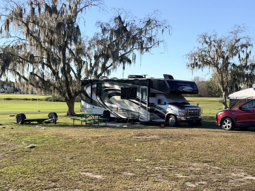 A Class C RV, specifically a Coachmen Leprechaun, is parked on a grassy campsite beside a red SUV and a small trailer, with moss-draped trees and open fields in the background. This setup showcases one of the popular types of RVs used for road trips and camping.