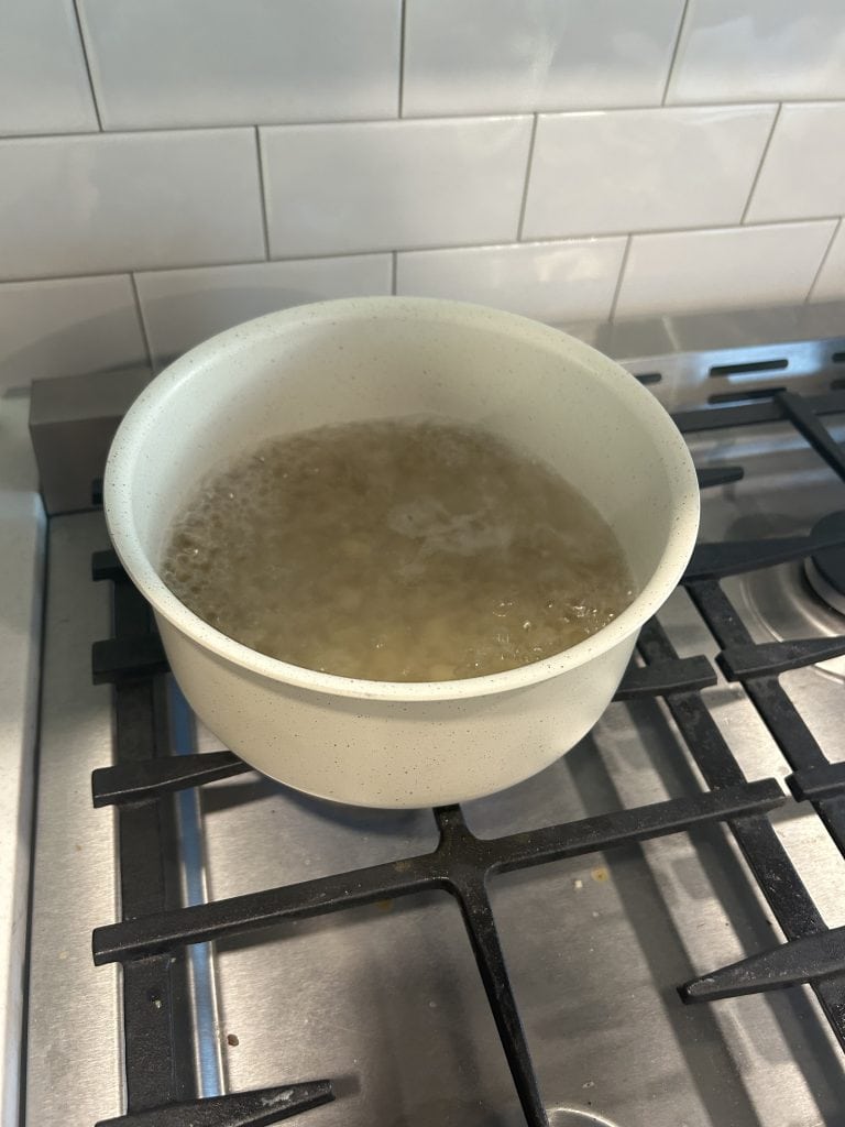 A cream-colored Gourmetific™ pot is shown on a gas stove with water at a rolling boil, set against a white tile backsplash. The image demonstrates the pot’s functionality for stovetop cooking.