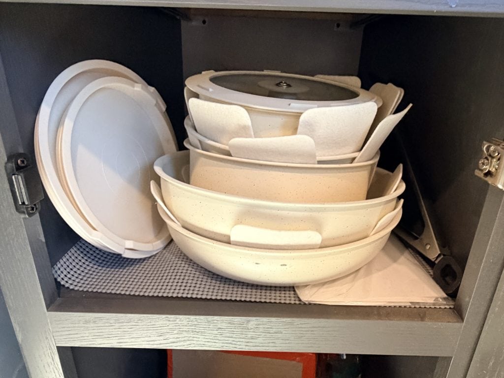 A stack of cream-colored Gourmetific™ cookware is neatly stored in a cabinet with protective felt pads between each piece. Matching lids are propped upright on the side, showcasing the set’s compact, organized storage design.