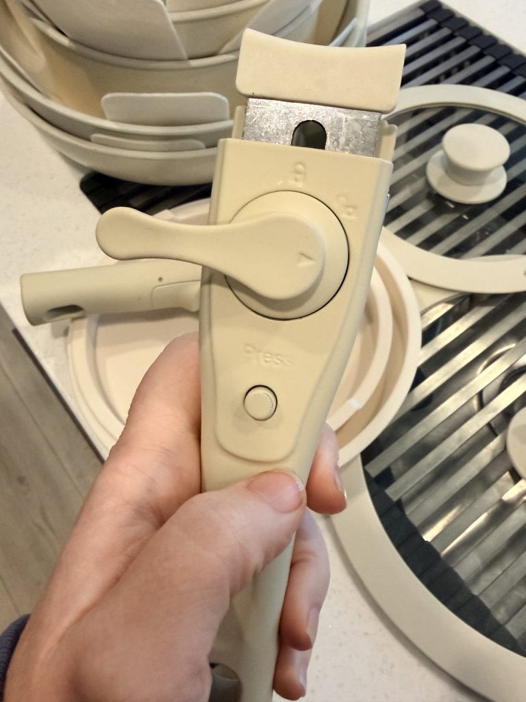 Close-up of a hand holding a cream-colored detachable handle from the Gourmetific™ Cookware Set, showing the locking mechanism and "Press" button. The stackable pots and matching lids are visible in the background, emphasizing the set's modular design.