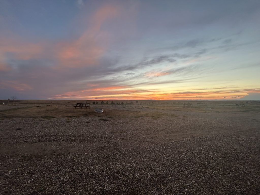 Wide open gravel and grassy RV campsite at sunset with scattered picnic tables and fire rings, set against a dramatic sky filled with soft pink, orange, and blue hues. The peaceful, expansive landscape captures the quiet beauty often found at rural Harvest Hosts locations.