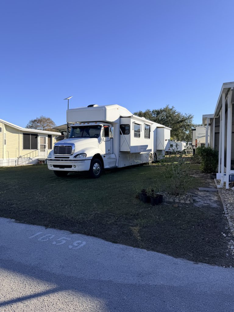 A large white Super C RV with extended slide-outs is parked on a grassy lot between mobile homes under a clear blue sky. This image illustrates one of the more heavy-duty types of RVs, built on a truck chassis for added power and space.