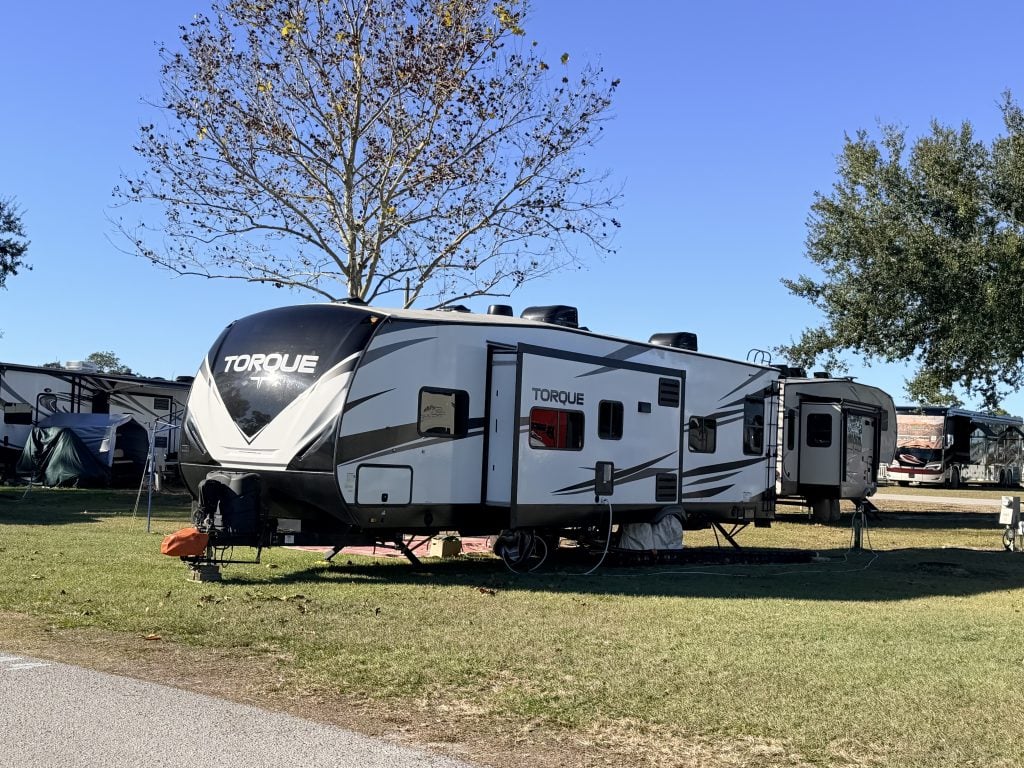 A Torque travel trailer toy hauler is set up on a grassy campsite with slide-outs extended, surrounded by other RVs and trees under a clear blue sky. This image represents one of the towable types of RVs designed for both living space and transporting recreational gear.