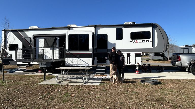 A couple stands with their dog in front of a Valor fifth-wheel toy hauler at a campground, complete with a side patio and connected pickup truck. This spacious setup exemplifies what to look for when you want to pick the right RV for comfort, versatility, and outdoor living.