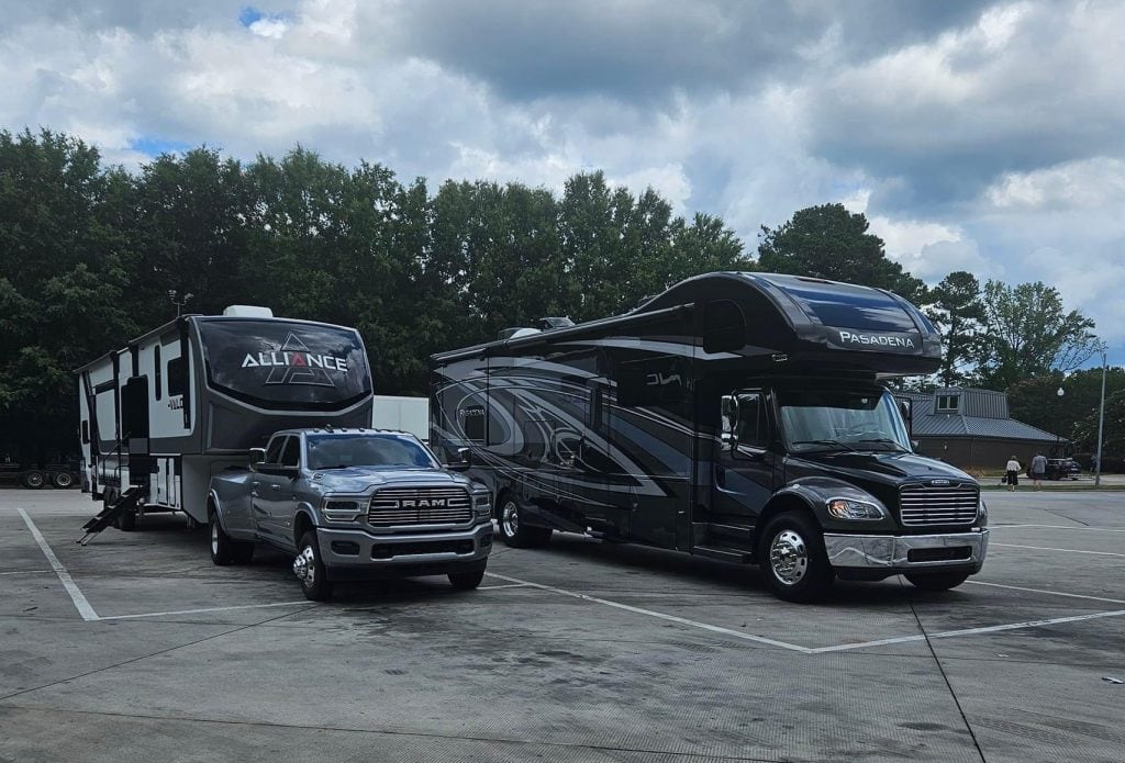 A fifth-wheel trailer hitched to a silver Ram truck is parked beside a large Super C RV labeled "Pasadena" in a spacious lot under a cloudy sky. This image highlights two distinct types of RVs, showcasing both towable and motorized options for travelers.