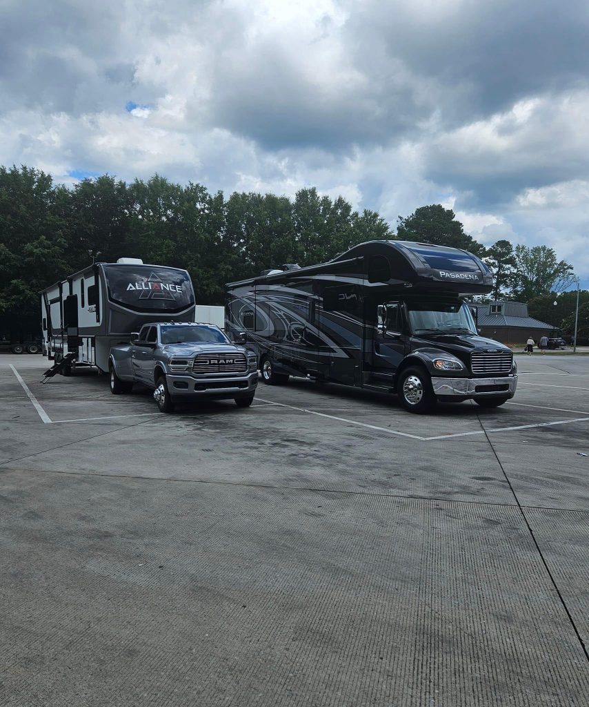 A fifth-wheel trailer hitched to a silver Ram truck is parked beside a large Super C RV labeled "Pasadena" in a spacious lot under a cloudy sky. This image highlights two distinct types of RVs, showcasing both towable and motorized options for travelers.