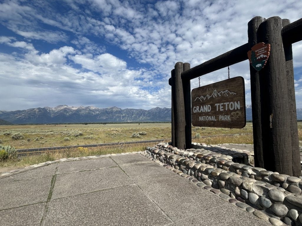 A rustic wooden sign reading “Grand Teton National Park” stands at the park entrance, backed by a sweeping view of open fields and the rugged Teton mountain range under a partly cloudy sky. The stone-lined base and wooden posts blend naturally with the scenic landscape.