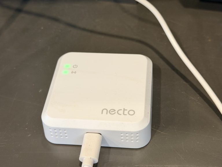 Close up of the necto RV pet temperature monitor plugged in on a countertop with green indicator lights showing power and connectivity. The small white device with a USB cable attached highlights the compact design featured in this necto RV pet temperature monitor review for monitoring RV conditions while pets are inside.